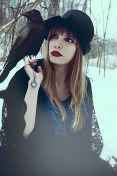 Photograph of a caucasian young woman with blonde hair and red lipstick holding a necklace in her left hand with a clock face on it. She is wearing vintage clothing and sitting in a woodlands area with a black crow perched on her right shoulder. TThe plac