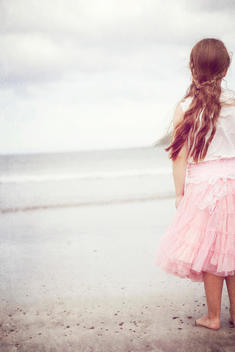 A young girl dressed in a pink sleeveless top and pink layered tulle skirt with long hair partially braided (plaited) stands looking at to the ocean at the waters edge of a beach. A pale blue sky, grey and white clouds, gently rolling waves. A textured ph