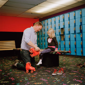 Chris, middle-aged father, kneels on ground to help his youngest daughter Mabel put on her roller skates at his boss\'s birthday party. Orem, Utah