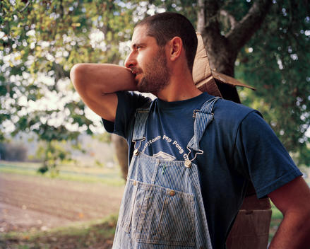 A young male farmer in overalls stands looking over his land, with one hand on his hip and the other over his shoulder holding a cardboard box.