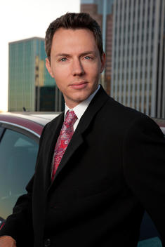 Corporate man in suit leaning on red car; city buildings in background