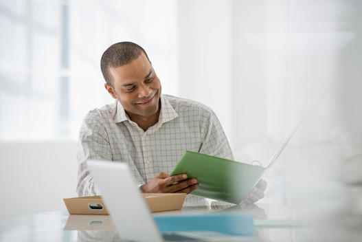 Business. A Man Seated At A Desk Opening A File Of Paperwork. A Laptop Open.