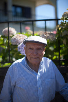 Older gentleman with a newsboy cap on in front of a garden.