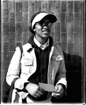 Student Of African American Appearance, Dressed In Sun Visor And Denim Jacket With Corn-Row Hair Style