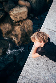 graphic down image of 6 year old blonde boy in black t-shirt laying on stone wall with rocky ocean below him.