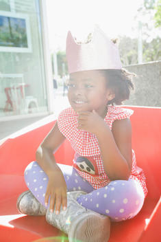 Smiling portrait of 6 year old girl African American girl wearing pink sparkle crown with hand on chin and bright sunlight.