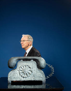 Dr. Irwin Jacobs, cofounder of Qualcomm, sitting behind a table with a huge metal sculpture of a rotary phone