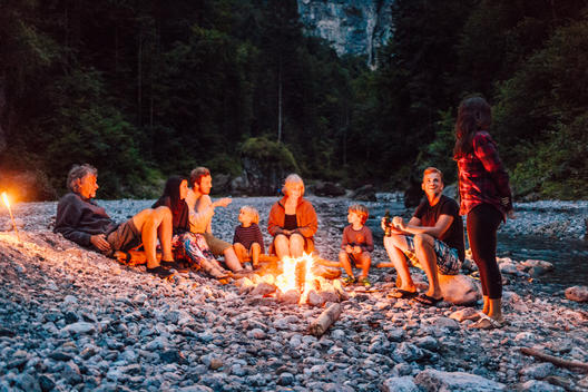 group of people of all ages sitting around campfire at rocky creek in mountainous landscape