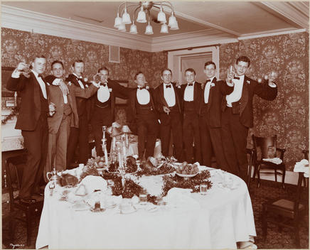 Men In Tuxedos Standing On Chairs Around A Set Dining Table, Raising Their Glasses In A Toast.