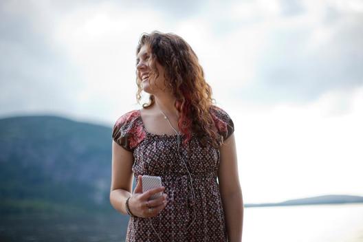 Portrait of young female caucasian college student listening to music with ear buds on mobile phone smiling while standing near misty river view