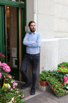 Young man with beard standing outside door with flower either side of him on pavement