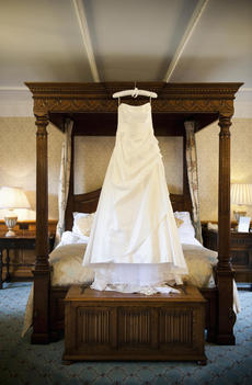 A full length white wedding dress hanging from a hanger on a four-poster bed.