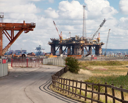 Industrial landscape featuring a wooden fenced road curving towards two large steel oil platforms,