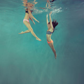 Two young women underwater, one swimming up with arms up and one reaching down with face out of water