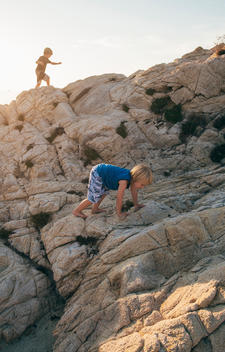 3 year old blonde boy in blue shirt climbing rocks with 6 year old blonde brother in background.
