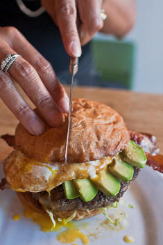 Woman’S Hand Cutting Into A Massy Hamburger With Fried Egg, Avocado, Lettuce, Bacon While Egg Yolkd Drips Out.