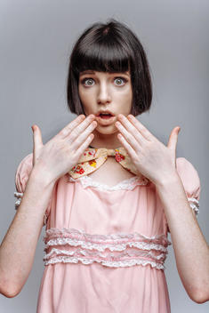 Cute young woman wearing old style pink dress and large bowtie. Short, dark haircut. Neutral background. Hands adjusting bowtie. Surprised expression.