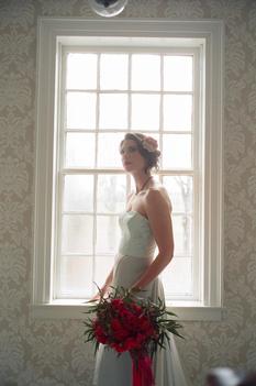 Bride waiting by window with red floral bouquet and large pattern wallpaper in old New England farmhouse.