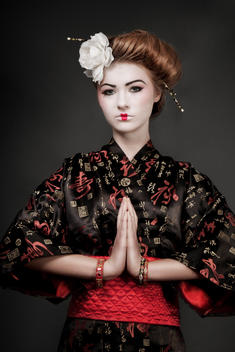 Studio beauty portrait brunette female model wearing a bun a white flower and chopsticks in her hair with traditional Japanese makeup in prayer position dressed in a kimono looking into camera