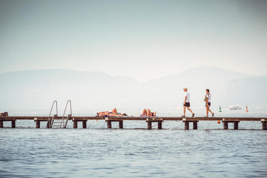 A couple walks on a wooden pier, some other people lay down and relax, blurred water in the foreground and blurred mountains in the background