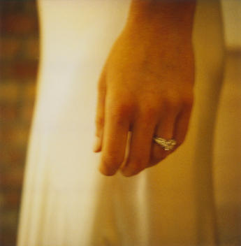 A close up polaroid of a bride's left hand, with her engagement ring, delicately resting next to her body in a white silk gown.