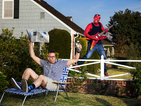 A man sitting in a lounge chair in his front yard spills his drink as his neighbor dressed as a devil jumps behind him playing a guitar