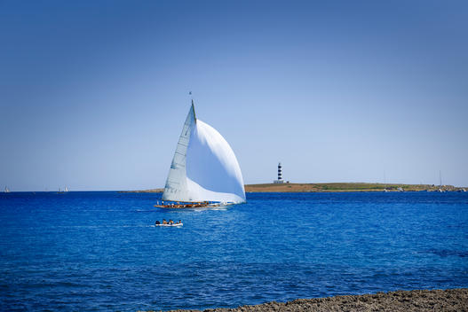 a yacht with a big white sail passing a lighthouse