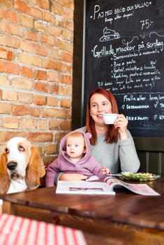 Happy young woman with baby girl with Basset Hound having coffee at cafe table