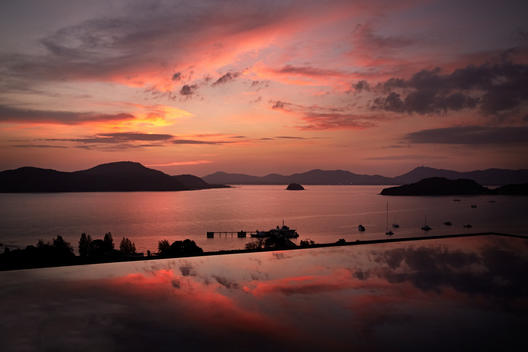 The evening sky is reflected in a swimming pool, as the silhouettes of mountains pierce through the sea and a small scattering of boats cluster by the beach