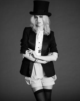 blonde fashion model in studio against a grey background, wearing a black tuxedo jacket, shorts and a tall black hat
