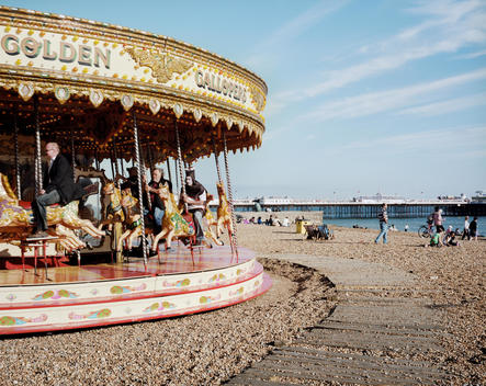 People enjoying a ride on a merry-go-round on Brighton beach. One man is dressed up in a costume. The Brighton Palace Pier can be seen in the background.