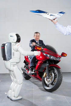 Honda CEO Takeo Fukui is sitting on a motorcycle with the ASIMO robot next to him and an arm holding a model plane above him