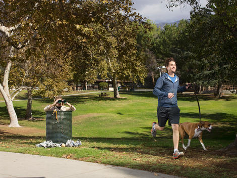 A man jogs in the park with his dog as a woman watches him with binoculars inside a trash can