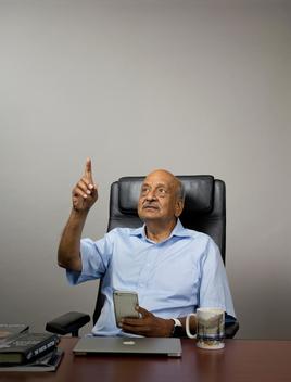 App inventor Ramesh Jain sits at his desk holding his smartphone and pointing his other hand up in the air