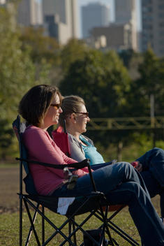 Two Mothers Sit In The Park And Chat While Their Sons Play Soccer.