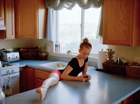 Portrait of a young blonde teenage girl wearing a ballerina outfit resting one leg up on the kitchen counter as she texts with friends on her cellphone in her family\'s suburban home. Pleasant Grove, Utah