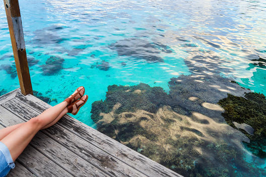 Overhead view of woman\'s legs, wearing shorts and sandals, lying on a dock, with clear blue water and a coral reeef below.