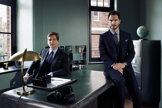 Two well suited men sit at 60\'s style desk in masculine retro office of a Savile Row tailors.