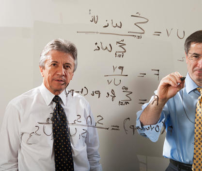 A businessman looks out through a glass wall as another man writes down math formulas with a marker