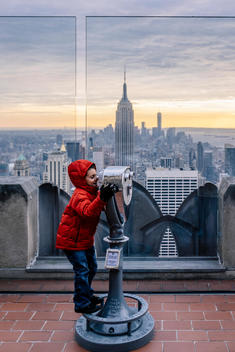 A boy looks through sightseeing binocular/telescope with the Empire State Building, One World Trade Center and lower Manhattan in the background at Top of the Rock at Rockefeller Center.