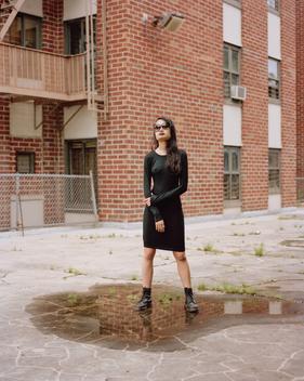 Fashion portrait of beautiful, disturbed Asian model/woman in black dress and sunglasses standing in puddle of water by housing projects in Brooklyn, NY