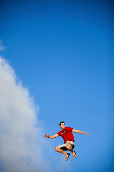 A young man in a red shirt jumping against a blue sky.