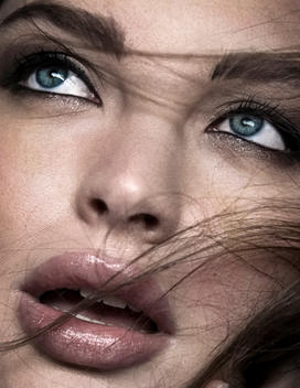Close up shot of Caucasian woman with wisps of brown hair streaming across the right side of her face. She has blue eyes.