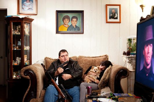 Man sitting on couch holding a gun watching TV as woman sleeps with her dog