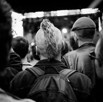 A man with a Mohawk and graying hair wearing a backpack stands in a crowd listening to famous 70s and 80s pop electronic band Devo at a music festival event near Times Square, NYC.