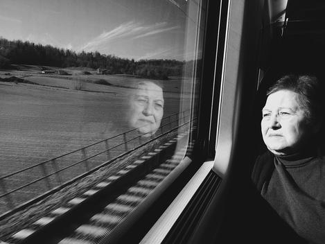 A older woman looks out of the train window with such sadness as you see her reflection in the window.