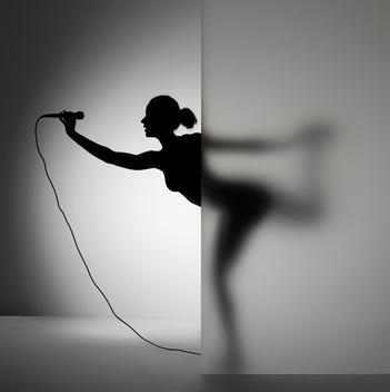 Dancer is half visible behind a frosted glass wall. She makes a nice figure - feminient. There is much ease in the image and radiates exclusivity and mental profit. She stands on one leg and holds up a microphone in front of her