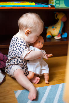 A baby in a polka dot onesie hugs a baby doll.
