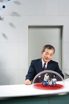 Honda CEO Takeo Fukui sits behind a desk with a glass globe containing a model ASIMO robot and motorcycle