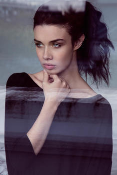 fashion model through reflective glass, with hands touching face, wearing a black sweater and hair in a ponytail
